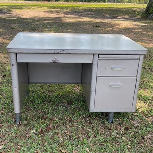 May include: A gray metal desk with a single drawer on the left side and two drawers on the right side. The desk has a simple design and is in good condition.