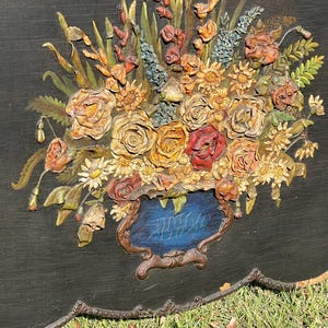 May include: A detailed, three-dimensional floral relief sculpture. The artwork features a bouquet of roses, daisies, and other flowers in shades of yellow, orange, and red, set against a black background. The flowers are arranged in a blue vase with a brown ornate frame.