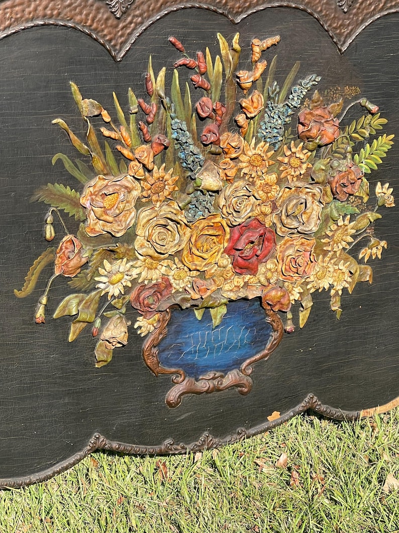 May include: A detailed, three-dimensional floral relief sculpture. The artwork features a bouquet of roses, daisies, and other flowers in shades of yellow, orange, and red, set against a black background. The flowers are arranged in a blue vase with a brown ornate frame.