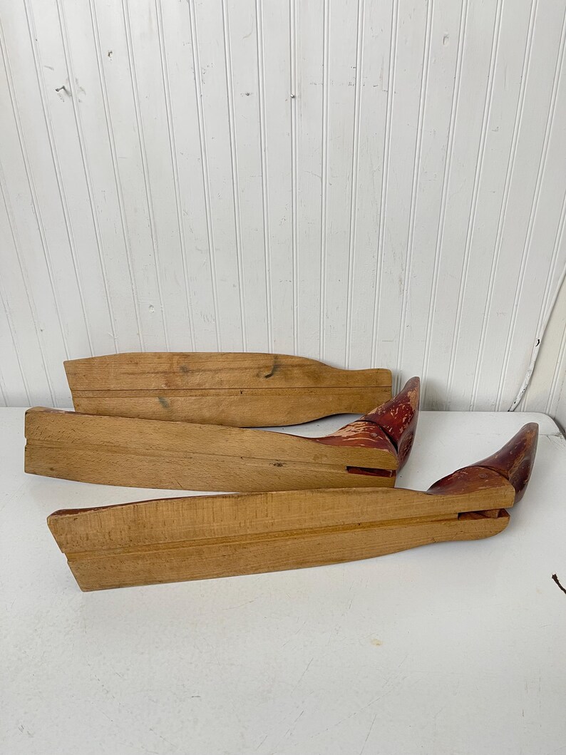 May include: Three wooden shoe forms, two are light brown and one is a darker brown with red paint. The forms are curved and have a rounded end.