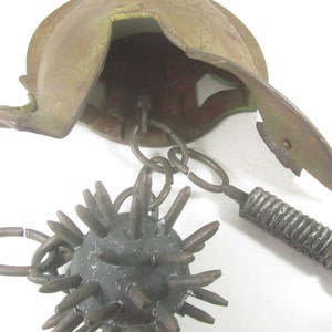 May include: A medieval-style spiked ball weapon with a chain attached to a helmet. The helmet is made of metal and has a rusty brown color. The spiked ball is made of metal and has a dark gray color.