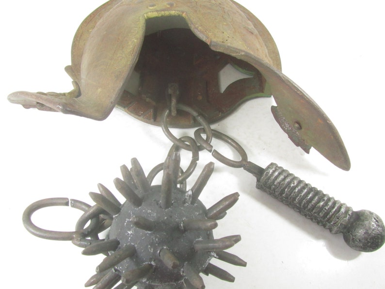 May include: A medieval-style spiked ball weapon with a chain attached to a helmet. The helmet is made of metal and has a rusty brown color. The spiked ball is made of metal and has a dark gray color.