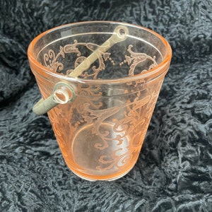 May include: A pink glass ice bucket with a silver handle and a delicate floral pattern. The bucket is empty and sitting on a black surface.