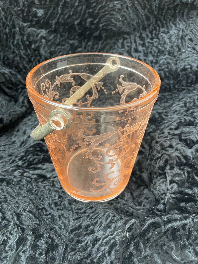 May include: A pink glass ice bucket with a silver handle and a delicate floral pattern. The bucket is empty and sitting on a black surface.