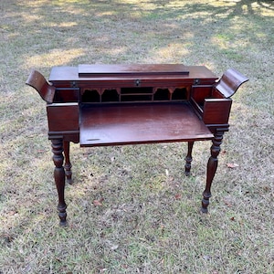 May include: A dark wood writing desk with a drop-down front and a series of small drawers. The desk has turned legs and is open, revealing the interior.