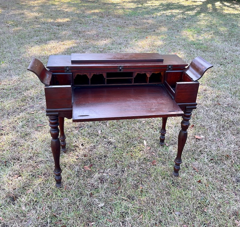 May include: A dark wood writing desk with a drop-down front and a series of small drawers. The desk has turned legs and is open, revealing the interior.
