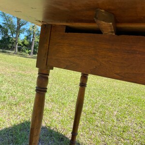 May include: A wooden table with a light brown tabletop and four turned legs. The table is set on a grassy surface, with the legs appearing to be approximately 28 inches tall. The table's construction is visible from underneath.
