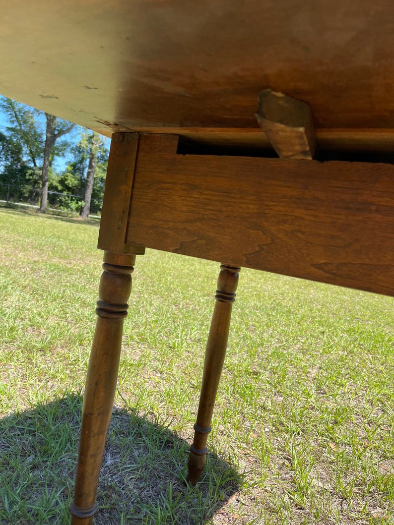 May include: A wooden table with a light brown tabletop and four turned legs. The table is set on a grassy surface, with the legs appearing to be approximately 28 inches tall. The table's construction is visible from underneath.