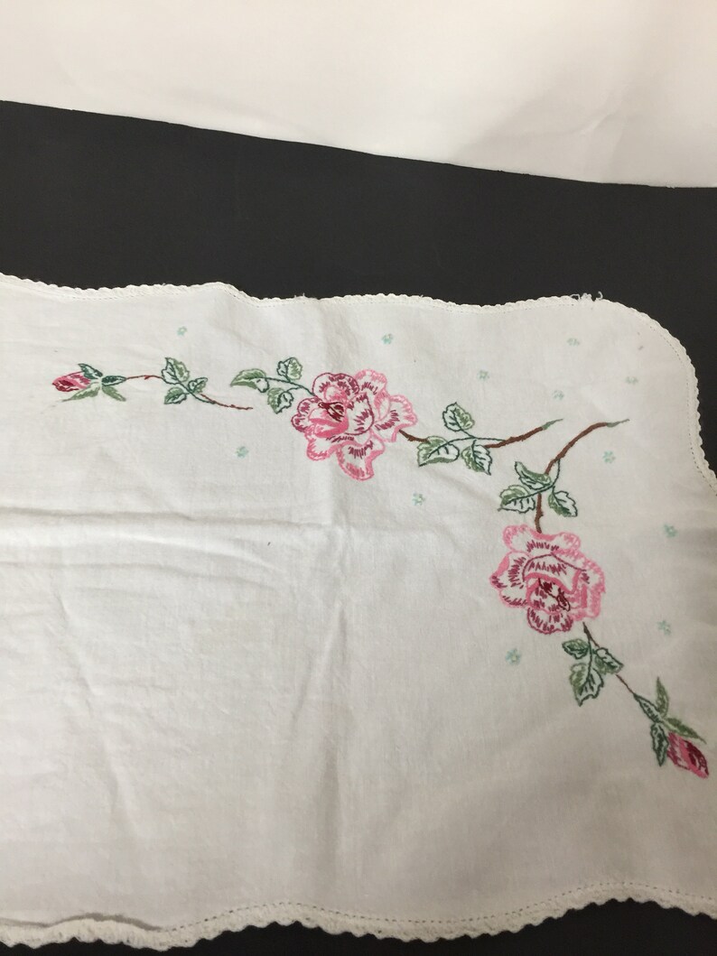 Op de afbeelding: Witte geborduurde tafelloper met roze rozen en groene bladeren. De loper heeft een gegolfde rand.