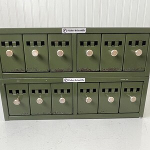 May include: A two-tiered, olive green metal cabinet with twelve small drawers. Each drawer has a silver knob and a rectangular vent. The cabinet has the "Fisher Scientific" logo on the top. The cabinet is likely used for storage.