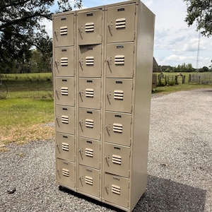 May include: A tall, beige metal locker unit with multiple individual compartments. Each compartment has a handle and ventilation slats. The lockers are outdoors on a gravel surface, with a backdrop of green grass, trees, and a blue sky.