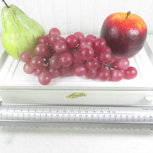 May include: White kitchen scale with a white tray and a silver measuring bar. The scale has a label that says "Anita". The tray has a green pear, a bunch of red grapes, and a red apple on it.