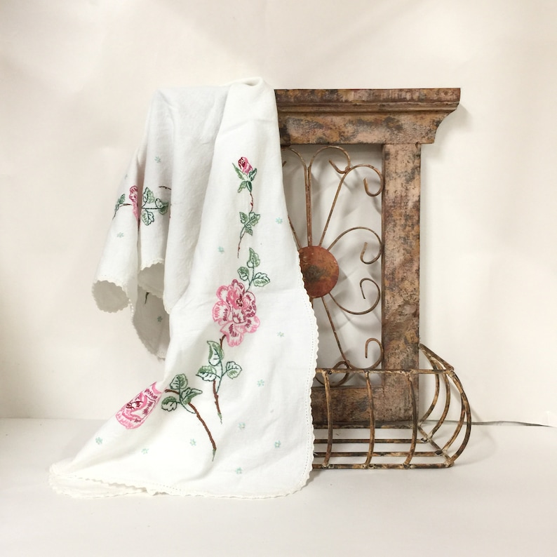 Op de afbeelding: Wit linnen tafelkleed met geborduurde roze en groene rozen. Het tafelkleed is gedrapeerd over een rustiek houten plank met een metalen frame.