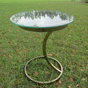 May include: A gold-toned side table with a round glass top. The table has a unique curved base.