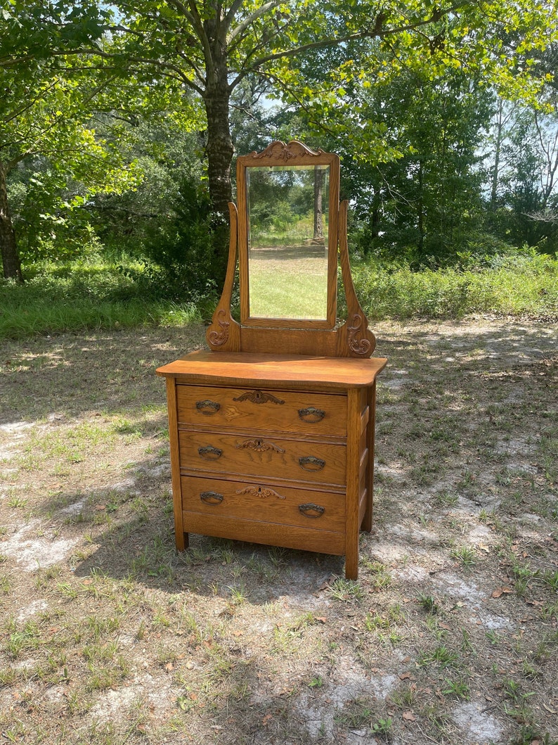 May include: Antique wooden dresser with a mirror. The dresser has three drawers with ornate metal pulls. The mirror has a carved wooden frame.