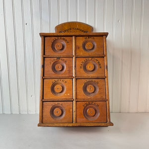 Könnte beinhalten: Ein antiker Gewürzschrank aus Holz mit acht Schubladen. Jede Schublade hat ein anderes Gewürzlabel, darunter Zimt, Macis, Piment, Backpulver, Nelken, Ingwer, Muskatnuss und Pfeffer.