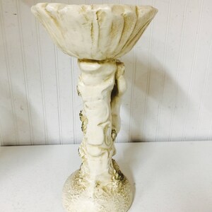 May include: A cream-colored, decorative pedestal bowl. The bowl has a scalloped edge and is supported by a sculpted figure. The base is round and textured. The overall design is ornate and vintage-inspired.