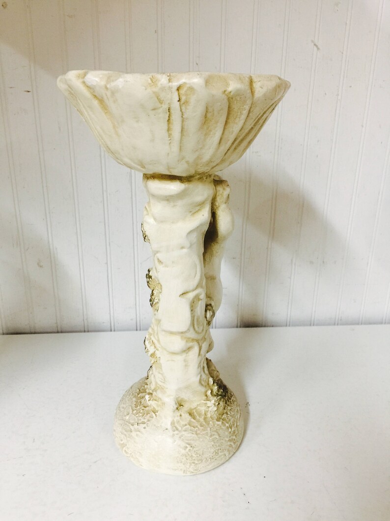 May include: A cream-colored, decorative pedestal bowl. The bowl has a scalloped edge and is supported by a sculpted figure. The base is round and textured. The overall design is ornate and vintage-inspired.