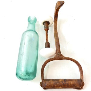 May include: An antique collection featuring a vintage teal glass bottle, a rusty metal and white porcelain door handle, and a rustic metal and wood stirrup. The items are arranged on a white surface.