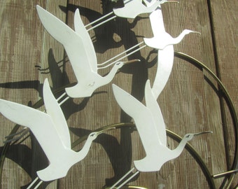 May include: White metal wall art featuring five stylized birds in flight. The birds are arranged in a circular pattern and are attached to a gold metal frame.