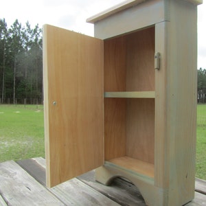 May include: A small wooden cabinet with a single door and a shelf inside. The cabinet is painted a light green color and has a distressed finish. The door is open and the inside of the cabinet is visible.