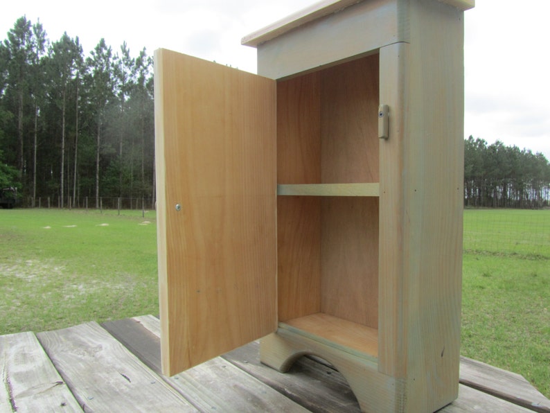 May include: A small wooden cabinet with a single door and a shelf inside. The cabinet is painted a light green color and has a distressed finish. The door is open and the inside of the cabinet is visible.