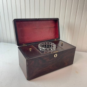 Puede incluir: Una caja de madera oscura con un inserto de vidrio y dos cajones pequeños. La caja tiene una cerradura y bisagras de latón.