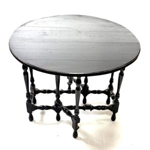 May include: A round, black wooden table with a dark finish. The table has a circular top and ornate, turned legs. The legs are connected by horizontal supports. The table is likely antique or vintage.
