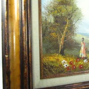May include: A framed oil painting depicting a woman in a pink dress standing in a field of wildflowers. The woman is looking to the right of the frame. The background features a lush green field, a river, and a forest with a large tree in the center.
