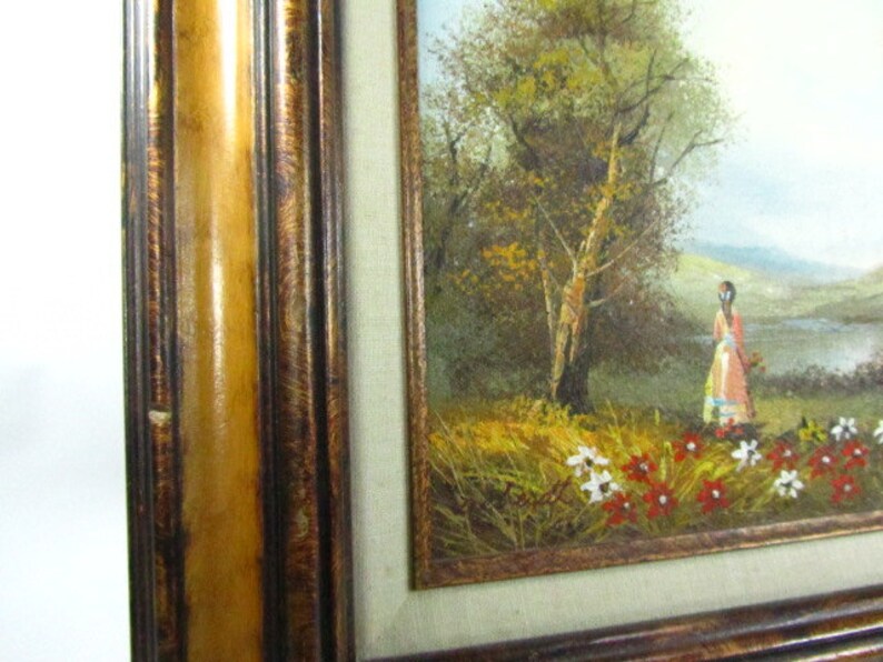 May include: A framed oil painting depicting a woman in a pink dress standing in a field of wildflowers. The woman is looking to the right of the frame. The background features a lush green field, a river, and a forest with a large tree in the center.