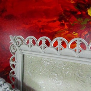 May include: A white ornate picture frame with a decorative, scalloped edge. The frame is sitting in front of a red, yellow, and orange abstract painting.