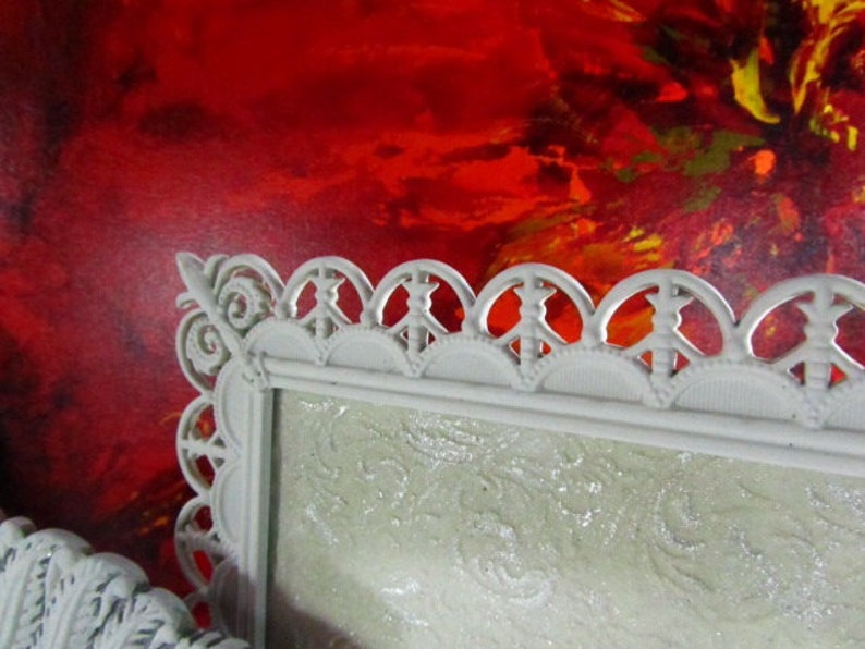 May include: A white ornate picture frame with a decorative, scalloped edge. The frame is sitting in front of a red, yellow, and orange abstract painting.