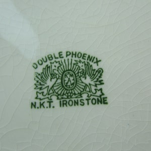 K&ouml;nnte beinhalten: Gr&uuml;nes und wei&szlig;es Double Phoenix N.K.T. Ironstone Logo mit zwei sich gegen&uuml;berstehenden Ph&ouml;nixv&ouml;geln.