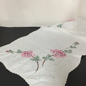 Op de afbeelding: Witte geborduurde tafelloper met roze rozen en groene bladeren. De loper heeft een gegolfde rand.