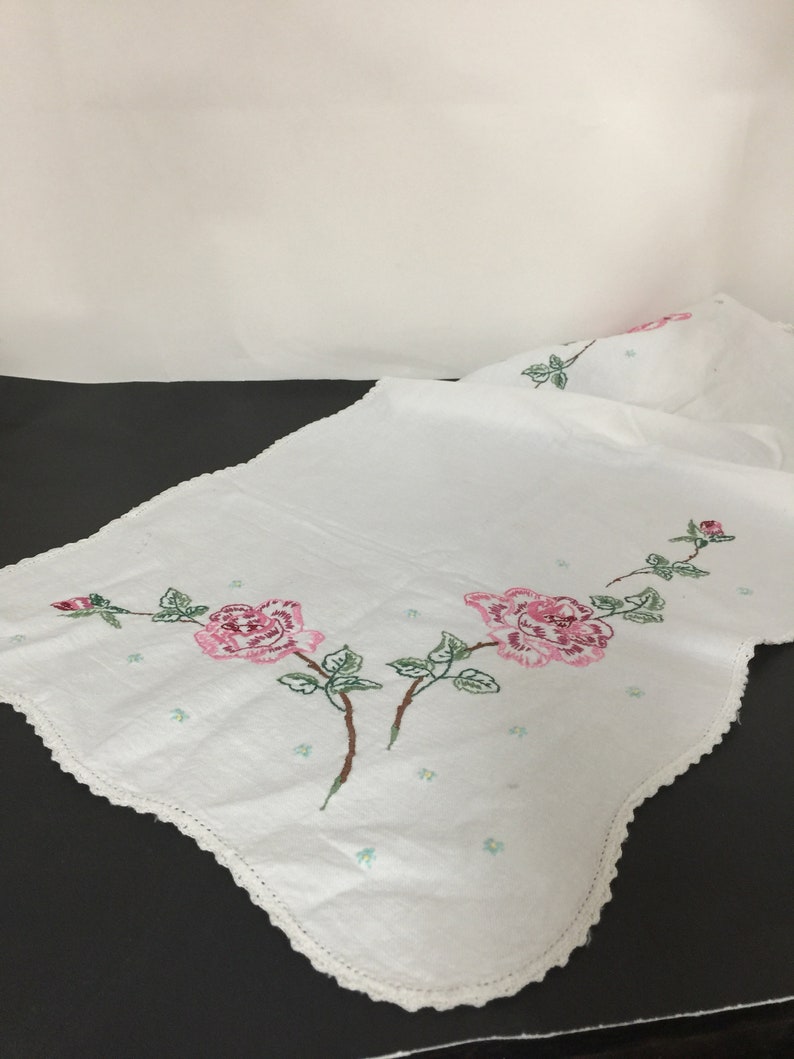 Op de afbeelding: Witte geborduurde tafelloper met roze rozen en groene bladeren. De loper heeft een gegolfde rand.