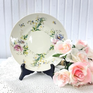 May include: Cream-colored ceramic plate with a floral design in shades of purple, blue, and yellow. The plate is displayed on a black stand, with a bouquet of pink roses in the foreground. The plate has a delicate, vintage aesthetic.