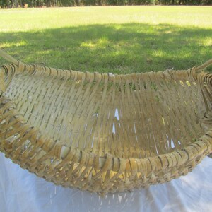 May include: A large, woven basket made of natural materials. The basket is oval shaped and has two handles. The basket is light brown in color and has a rustic appearance.