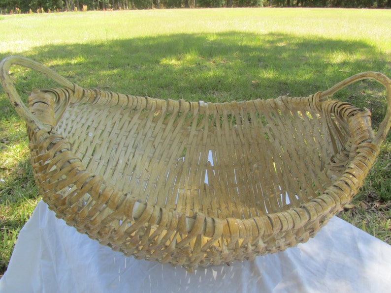 May include: A large, woven basket made of natural materials. The basket is oval shaped and has two handles. The basket is light brown in color and has a rustic appearance.