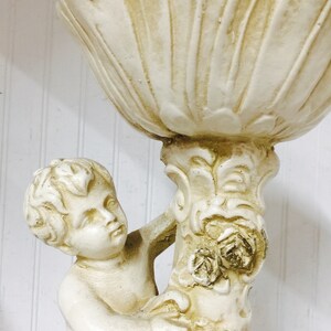 May include: Off-white cherub statue holding a large, shell-shaped bowl. The statue has a detailed design with floral accents on the column. The cherub's face is serene, and the overall aesthetic is classical.