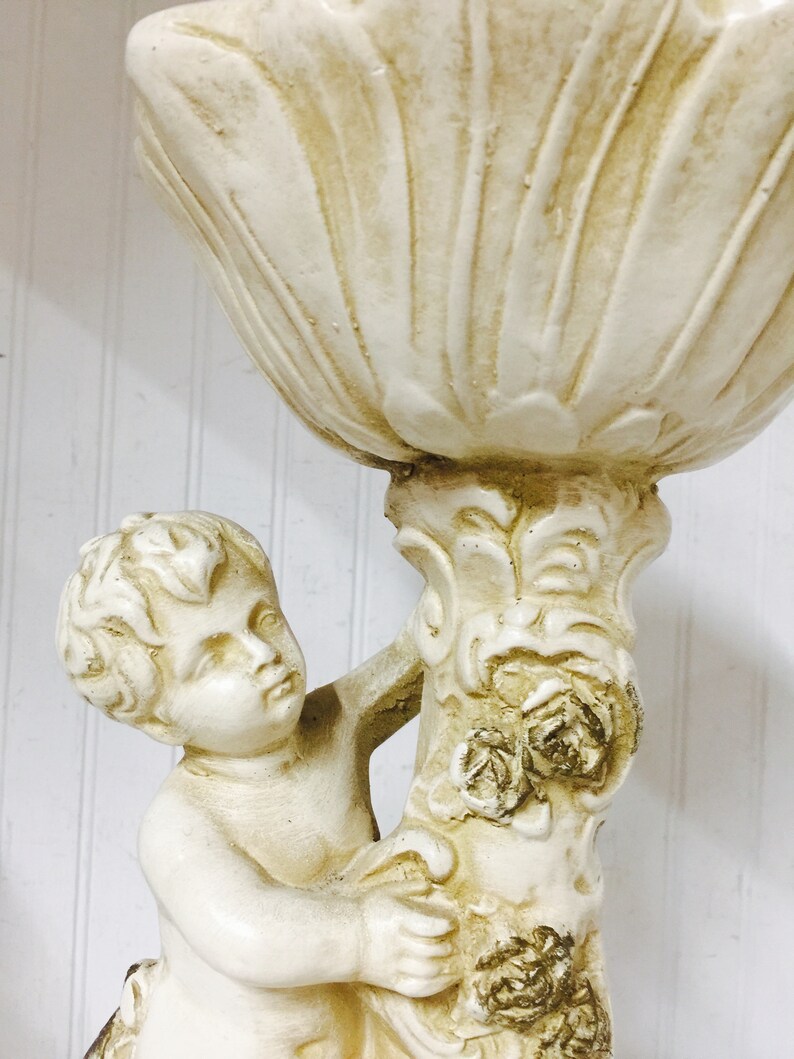 May include: Off-white cherub statue holding a large, shell-shaped bowl. The statue has a detailed design with floral accents on the column. The cherub's face is serene, and the overall aesthetic is classical.