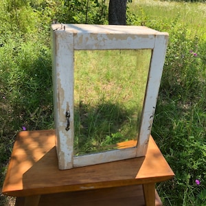 May include: A white wooden medicine cabinet with a mirror door and a black metal handle. The cabinet is sitting on a wooden table.