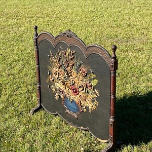 May include: An antique fireplace screen with a dark green background and a detailed floral arrangement in a blue vase. The screen has a decorative wooden frame with ornate carvings and stands on four legs.