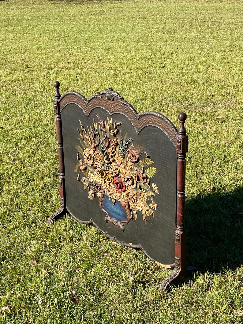 May include: An antique fireplace screen with a dark green background and a detailed floral arrangement in a blue vase. The screen has a decorative wooden frame with ornate carvings and stands on four legs.