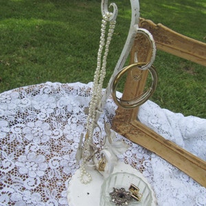 May include: White metal jewelry stand with a decorative scroll design. The stand has a small dish for rings and a hook for necklaces. A strand of pearls hangs from the hook.
