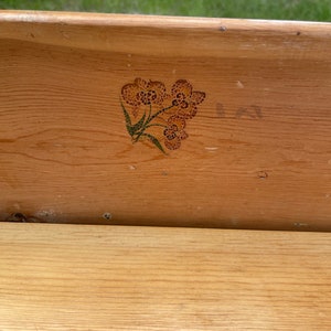 K&ouml;nnte beinhalten: Eine Holzkiste mit einem gemalten Blumendesign auf der Vorderseite. Das Design ist in Braun- und Gr&uuml;nt&ouml;nen gehalten.