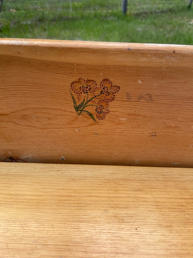 K&ouml;nnte beinhalten: Eine Holzkiste mit einem gemalten Blumendesign auf der Vorderseite. Das Design ist in Braun- und Gr&uuml;nt&ouml;nen gehalten.