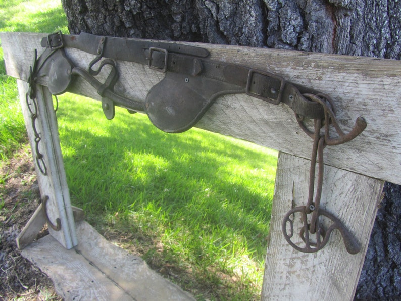 K&ouml;nnte beinhalten: Ein rustikaler Holzrahmen mit einem Lederriemen und Metallbeschl&auml;gen. Der Rahmen ist verwittert und hat eine nat&uuml;rliche Holzoptik. Der Riemen ist braun und hat eine Schnalle. Die Beschl&auml;ge sind aus Metall und rostig.