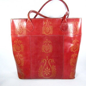 May include: A red leather tote bag with a gold paisley pattern. The bag has two top handles and a zipper closure.