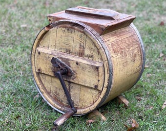 May include: Antique wooden butter churn with a hand crank. The churn is painted yellow and has a wooden lid. It is sitting on a grassy surface.