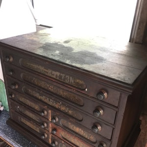May include: A dark wooden antique drawer cabinet with eight drawers. Each drawer has a glass front and a metal knob. The drawers are labeled with faded text, including "Cotton", "Dark", and "Arc".